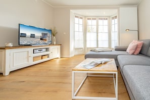 Un salon avec un téléviseur posé sur un meuble en bois, un canapé gris, une table basse en bois et une fenêtre avec des stores.