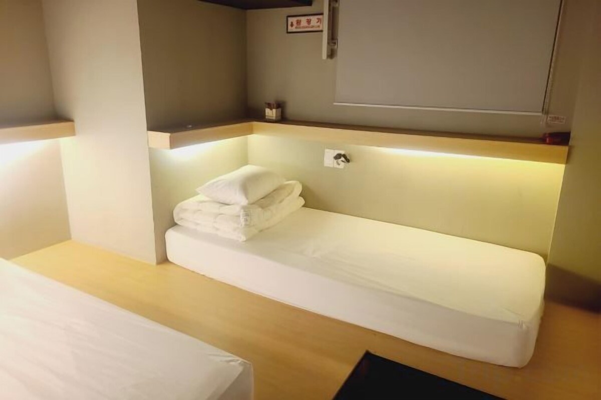 A compact hotel room with a single bed, a small bedside table, and a built-in bench with a pillow.