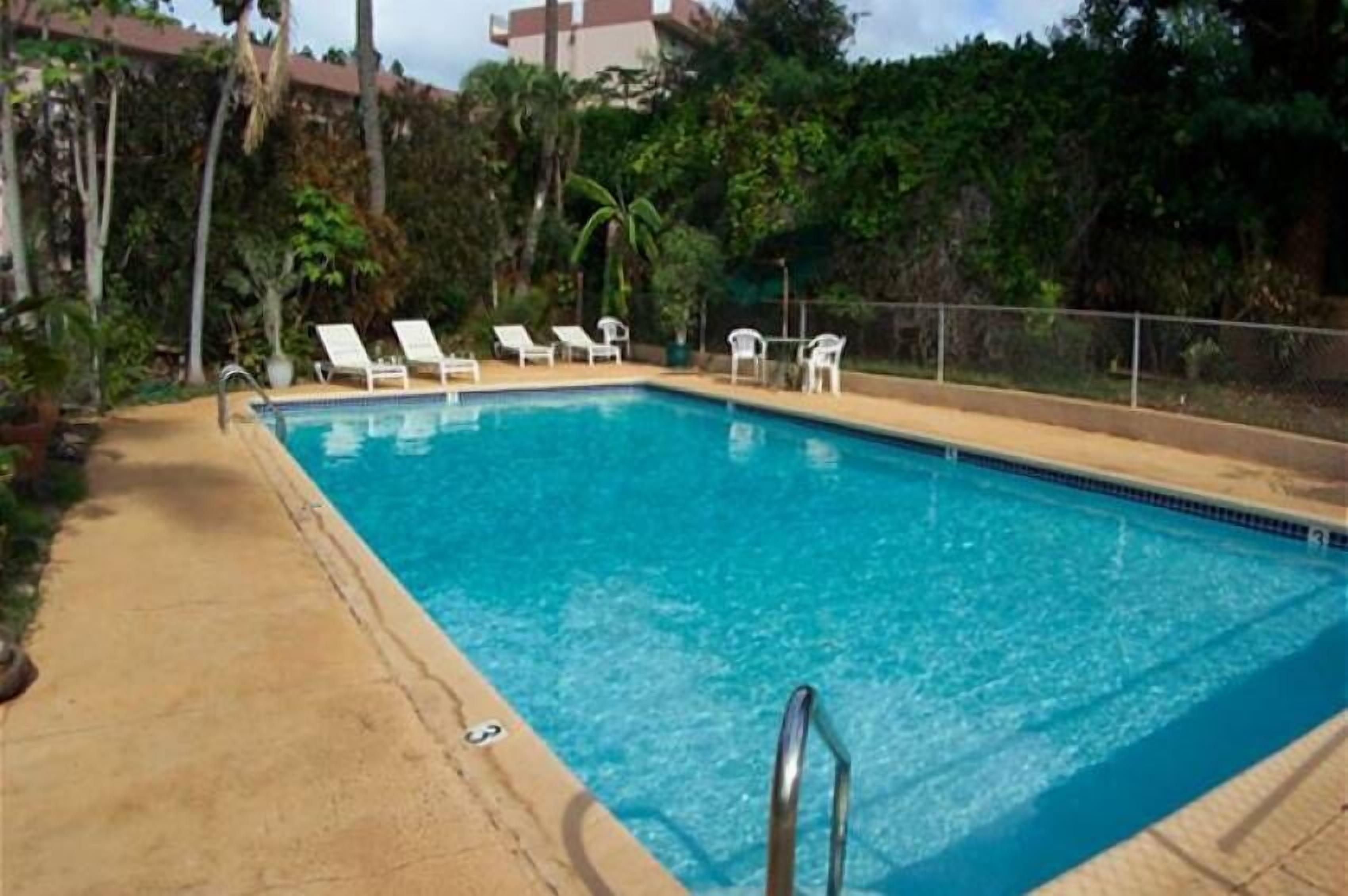 A rectangular swimming pool with clear blue water, surrounded by a concrete deck. There are several white lounge chairs aligned along the pool's edge.