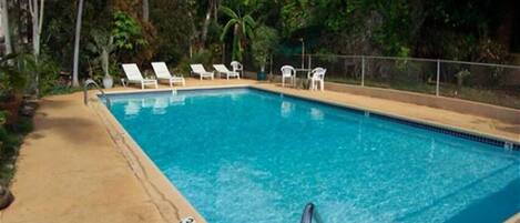 A rectangular swimming pool with clear blue water, surrounded by a concrete deck. There are several white lounge chairs aligned along the pool's edge.