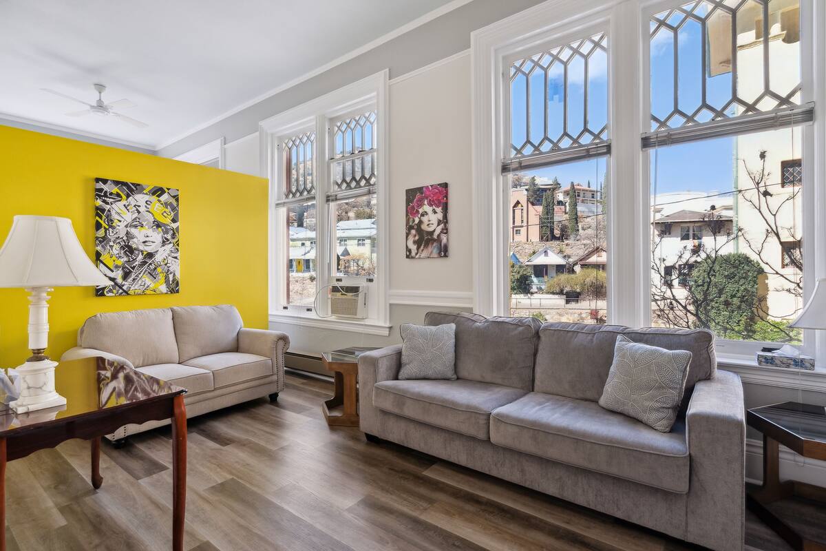 A living room with a yellow wall, two sofas, a table with a lamp, and large windows with a view of the neighborhood.