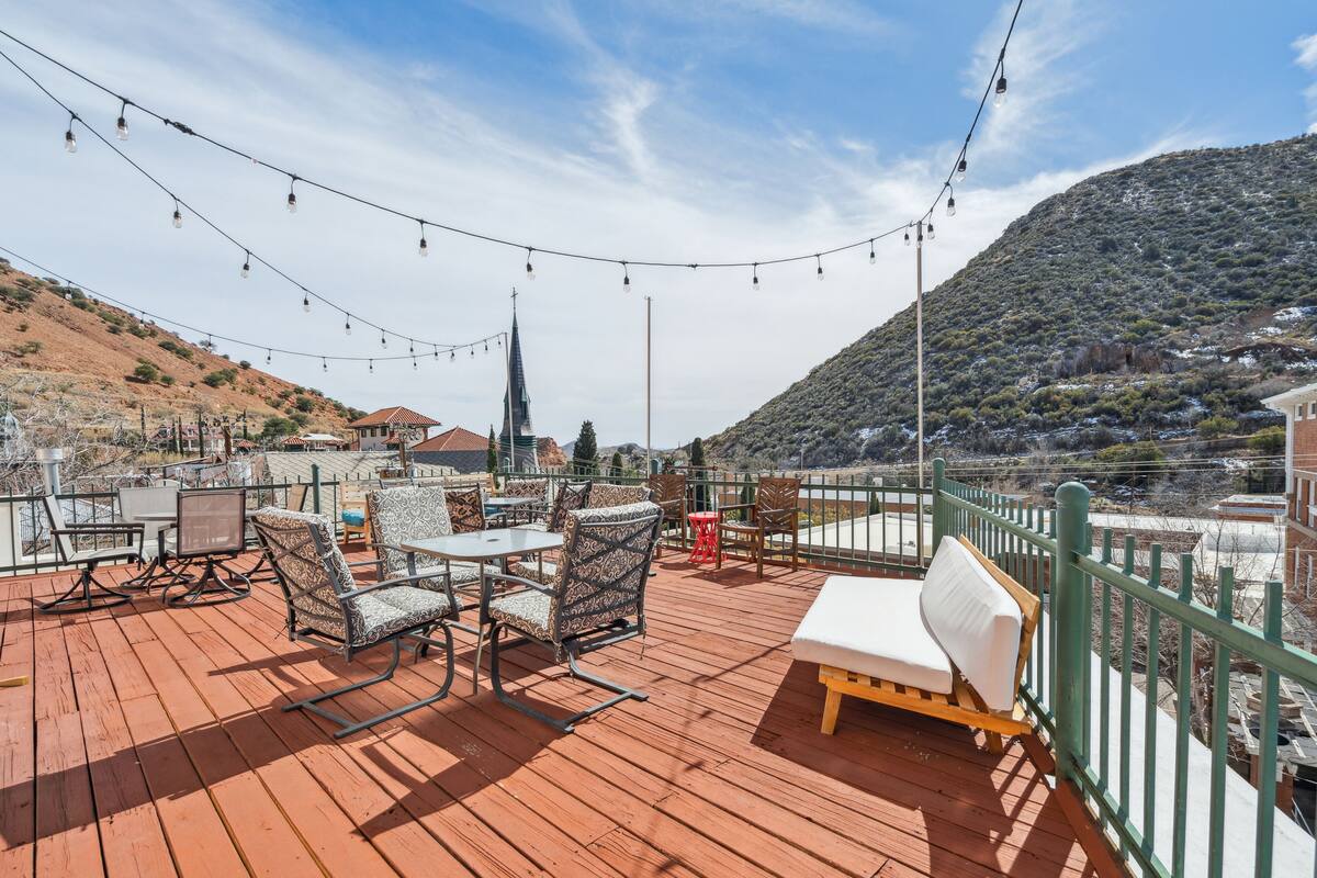 A wooden deck with outdoor seating, string lights, and a view of a hill and buildings.