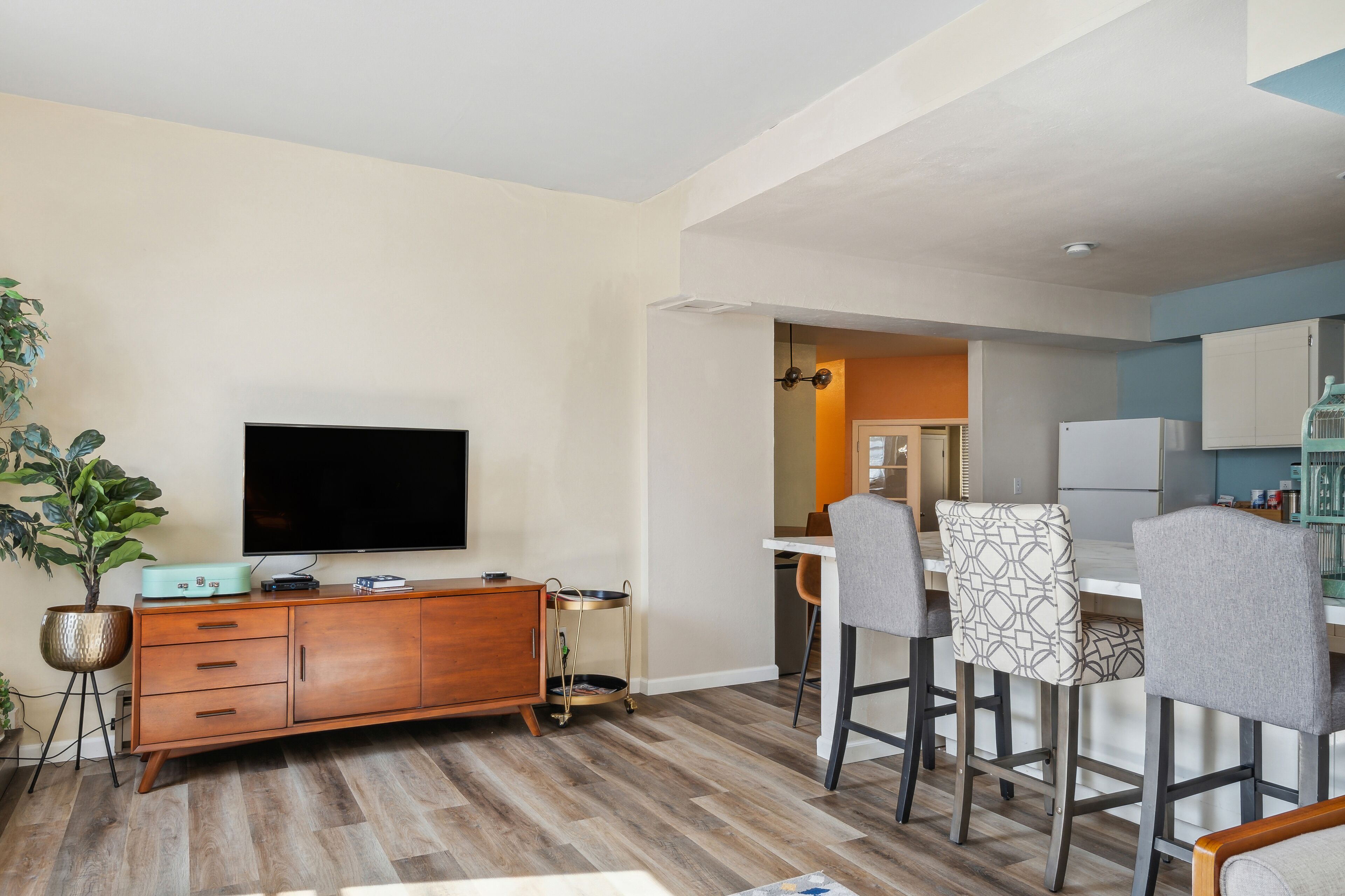 Een moderne woonkamer met een flatscreen televisie, een houten kast en een eethoek met barstoelen.