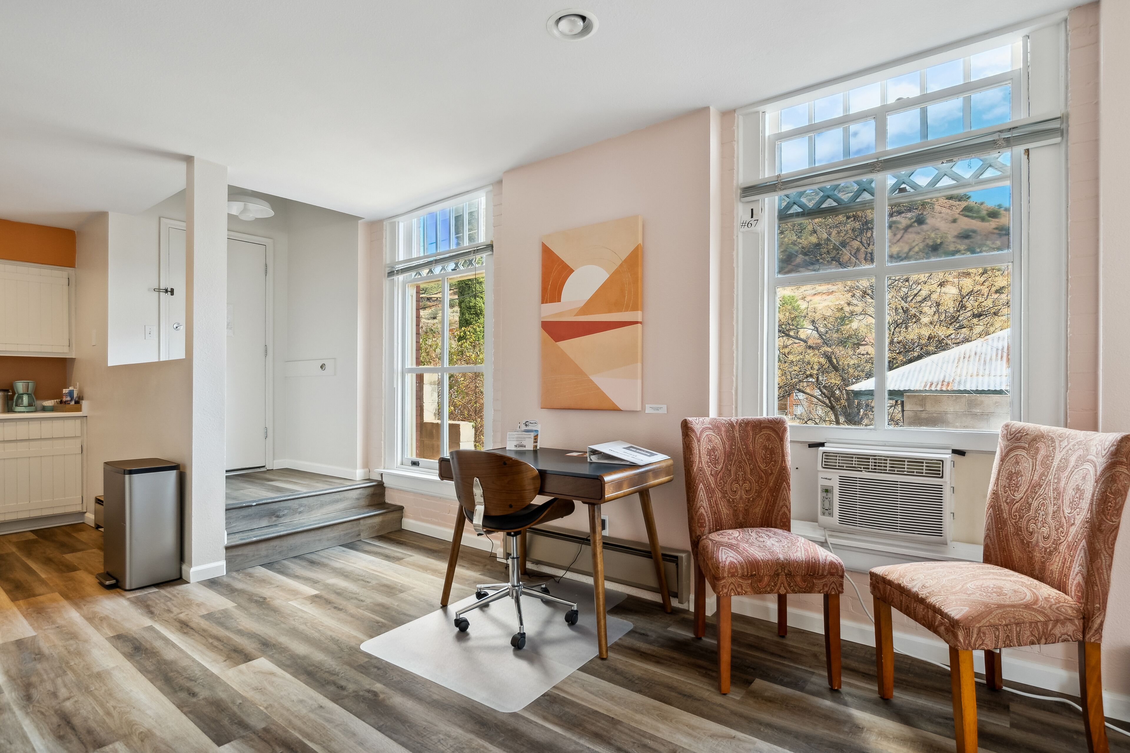 Una sala de estar moderna con un escritorio, dos sillas estampadas, un ventanal con vistas y una zona de cocina al fondo.