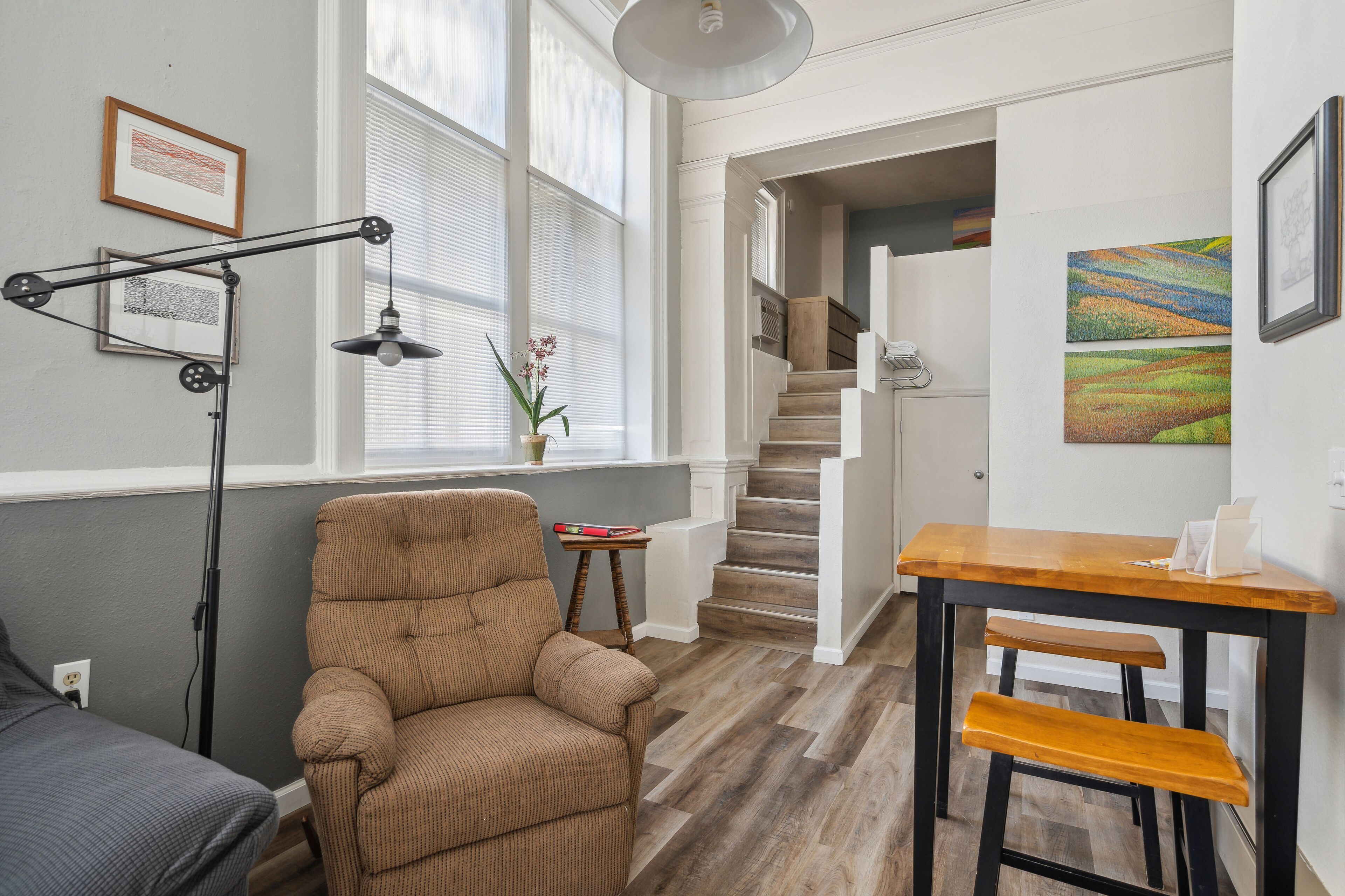 A room with a brown armchair, a wooden table, a floor lamp, and a staircase leading to another room.