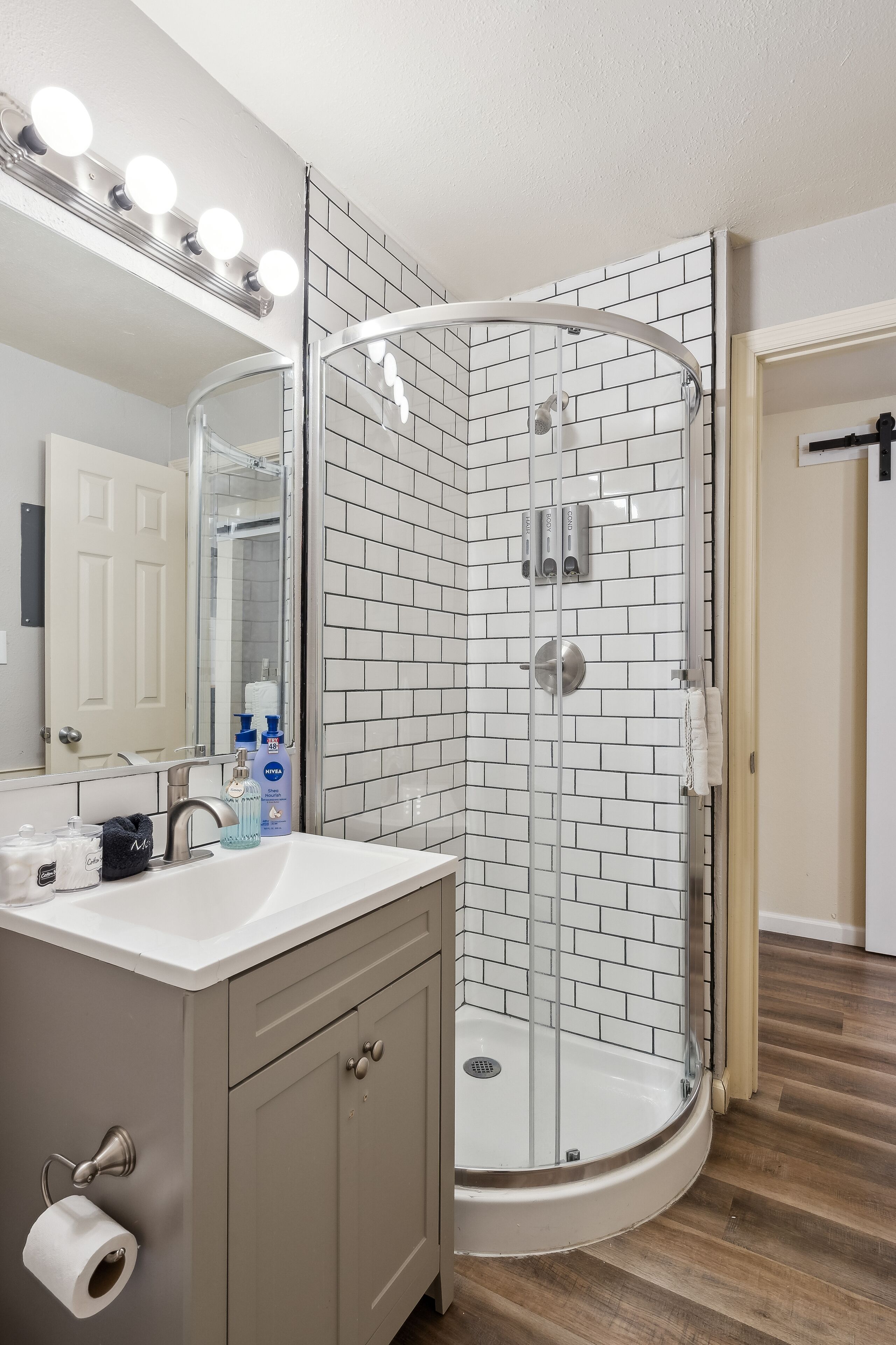 A modern bathroom with a glass-enclosed shower, white tiled walls, a vanity with a mirror, and a toilet paper holder.