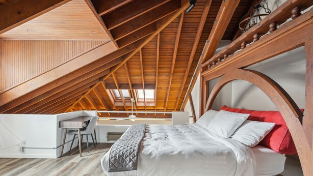 Ein Schlafzimmer mit schräg verlaufender Decke, einem Bettgestell aus Holz, einem Schreibtisch und einem Stuhl.
