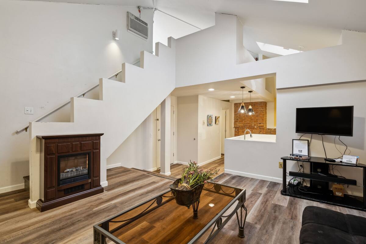 A modern living room with a fireplace, a coffee table with a plant, a staircase, a TV, and a kitchen area.