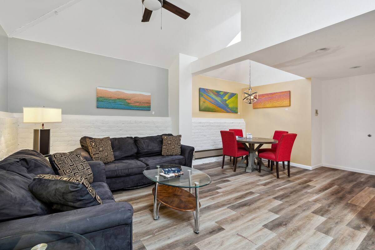 A modern living room with a glass coffee table, a dark sofa with patterned cushions, a red dining chair, and a painting on the wall.