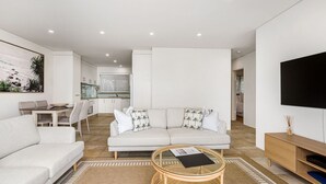 A modern living room with a sofa, a coffee table, and a TV. There is a dining area with a table and chairs. - Beach Break - Embrace the coastal lifestyle, with a view that will steal your breath away (East Ballina)