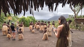 A group of people in traditional attire under a thatched roof, with a mountainous landscape in the background.