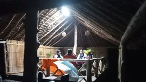 An interior view of a rustic hut with a thatched roof, a central table with chairs, and a hanging light fixture.