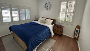 Een slaapkamer met een bed, twee nachtkastjes, een wandklok en twee ramen met jaloezieën.
