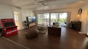 Een ruime woonkamer met een plafondventilator, een televisie, een gameconsole en uitzicht op de omgeving door grote ramen.