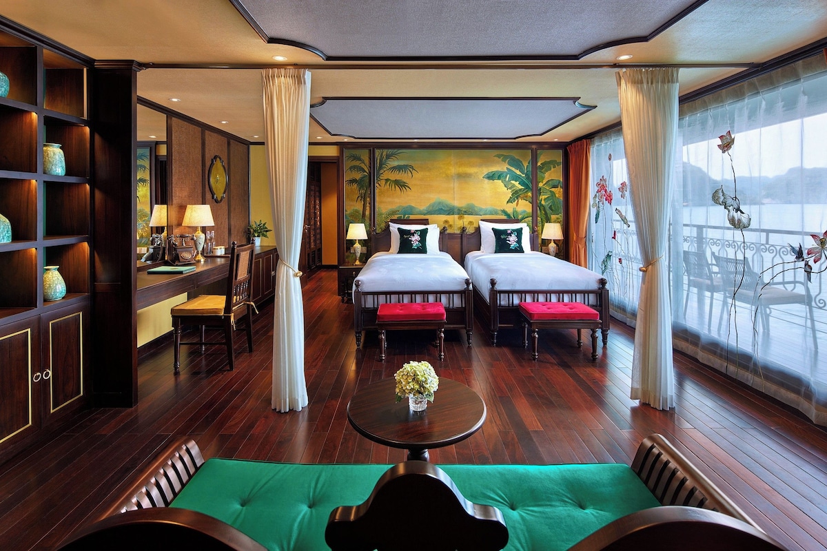 Une chambre d’hôtel avec deux lits, une table à manger, un canapé vert, une bibliothèque et une grande fenêtre donnant sur un beau paysage.