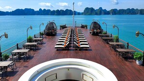 A deck with seating and tables, overlooking a body of water with distant landforms. - Indochine Premium Halong Bay (Tuan Chau)
