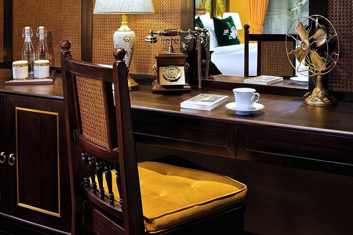 Une chambre d’hôtel avec un bureau en bois, une chaise avec un coussin jaune, une lampe, un ventilateur et un grand miroir qui reflète un lit avec un rideau jaune.