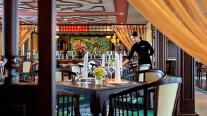 A restaurant with tables set for dining, a waiter attending to a table, and a decorative ceiling. - Indochine Premium Halong Bay (Tuan Chau)