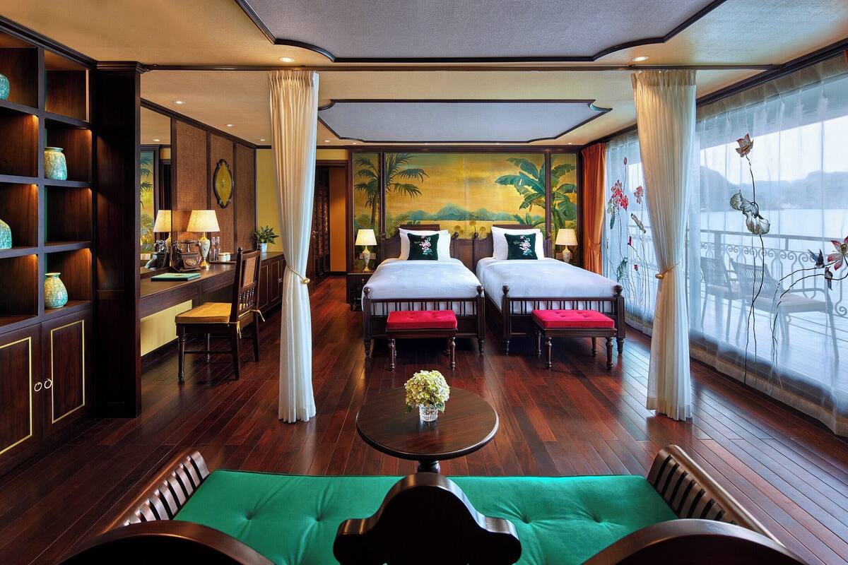 Een hotelkamer met twee bedden, een eettafel, een groene bank, een boekenkast en een groot raam met uitzicht.
