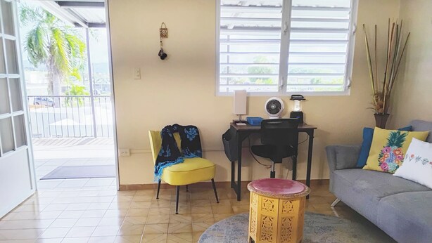 Un salón con una butaca amarilla, un sofá gris, un taburete de madera, un escritorio con un ventilador y una ventana con persianas.