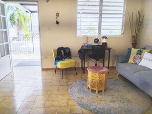 A living room with a yellow chair, a grey sofa, a small wooden stool, a desk with a fan, and a window with blinds. - Caguas Villa Santa Juana 2 Fully Equipped 2BR Home with SOLAR PANELS /A/C / WIFI (Caguas)