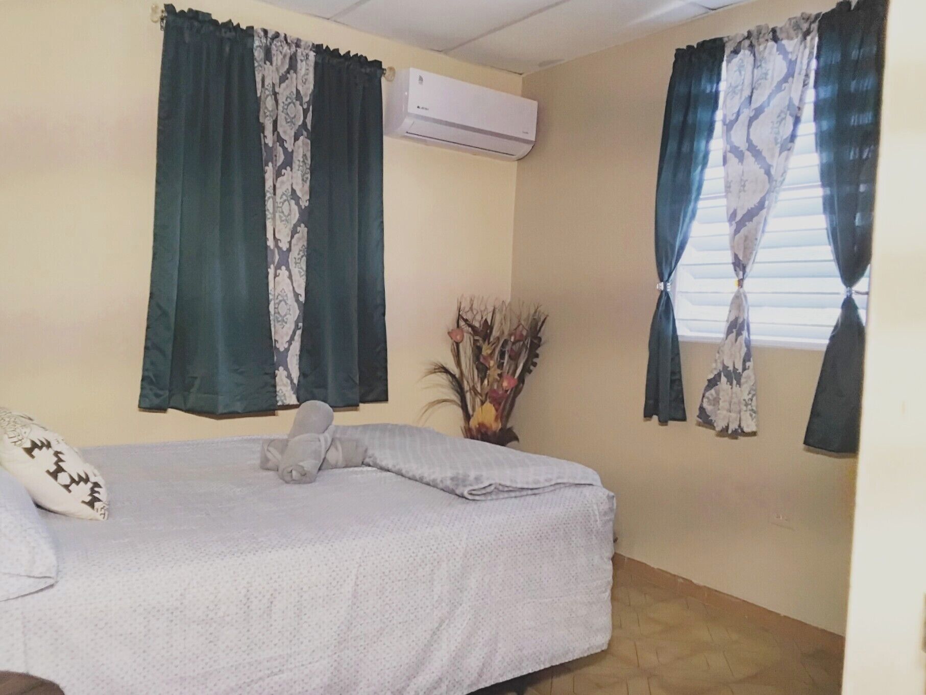Une chambre à coucher avec un lit, deux fenêtres avec des rideaux, un climatiseur et une plante décorative.