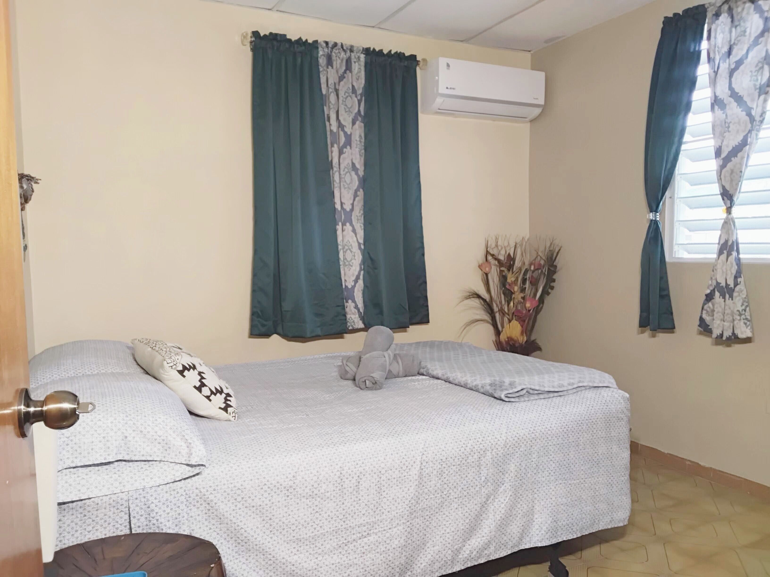 A bedroom with a bed, a decorative pillow, a wall-mounted air conditioner, and a window with curtains.