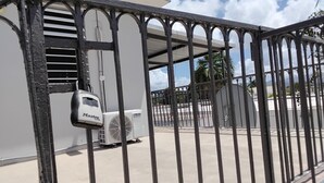Un área cercada con un candado de seguridad, un equipo de aire acondicionado y una vista despejada de un estacionamiento con palmeras y un edificio al fondo.