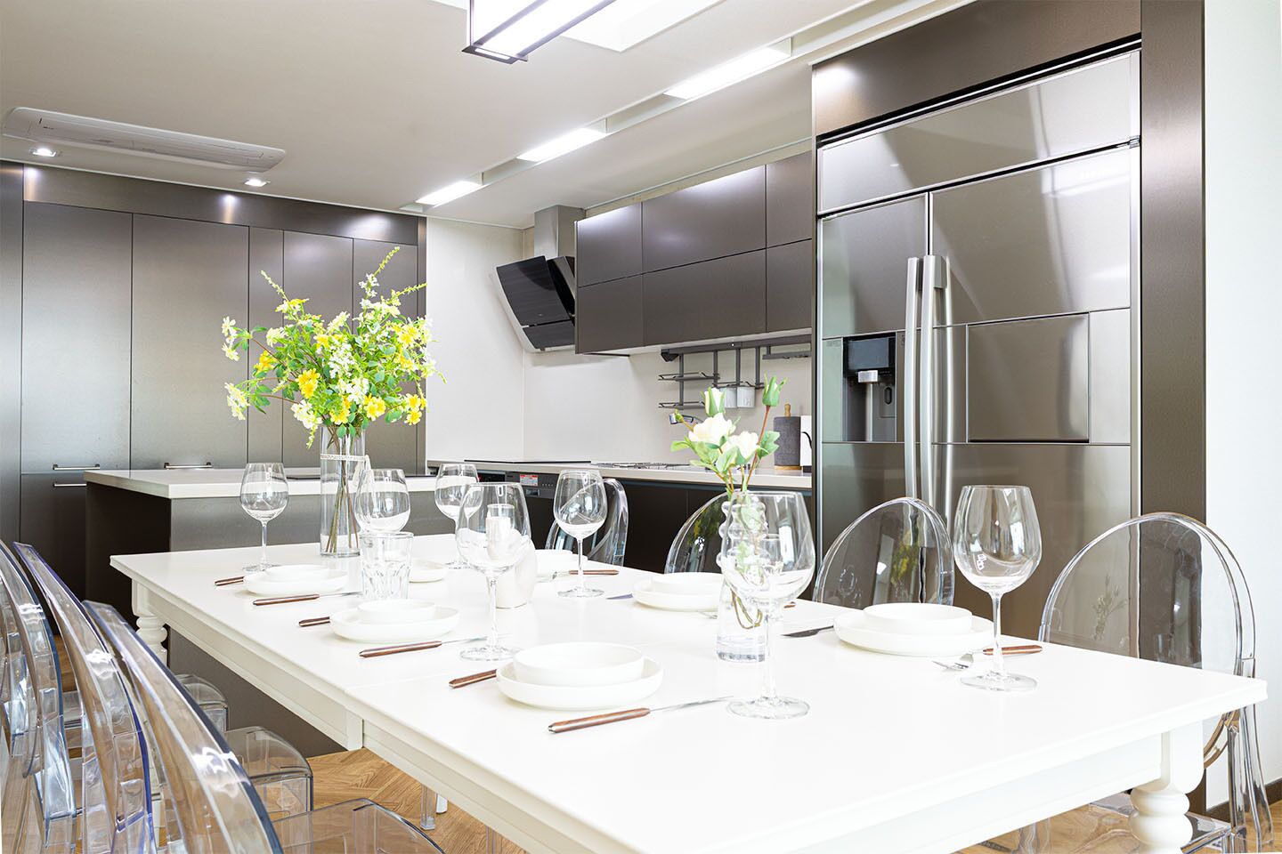 A modern kitchen with a long dining table set for six, equipped with stainless steel appliances, clear glassware, and a vase with flowers.