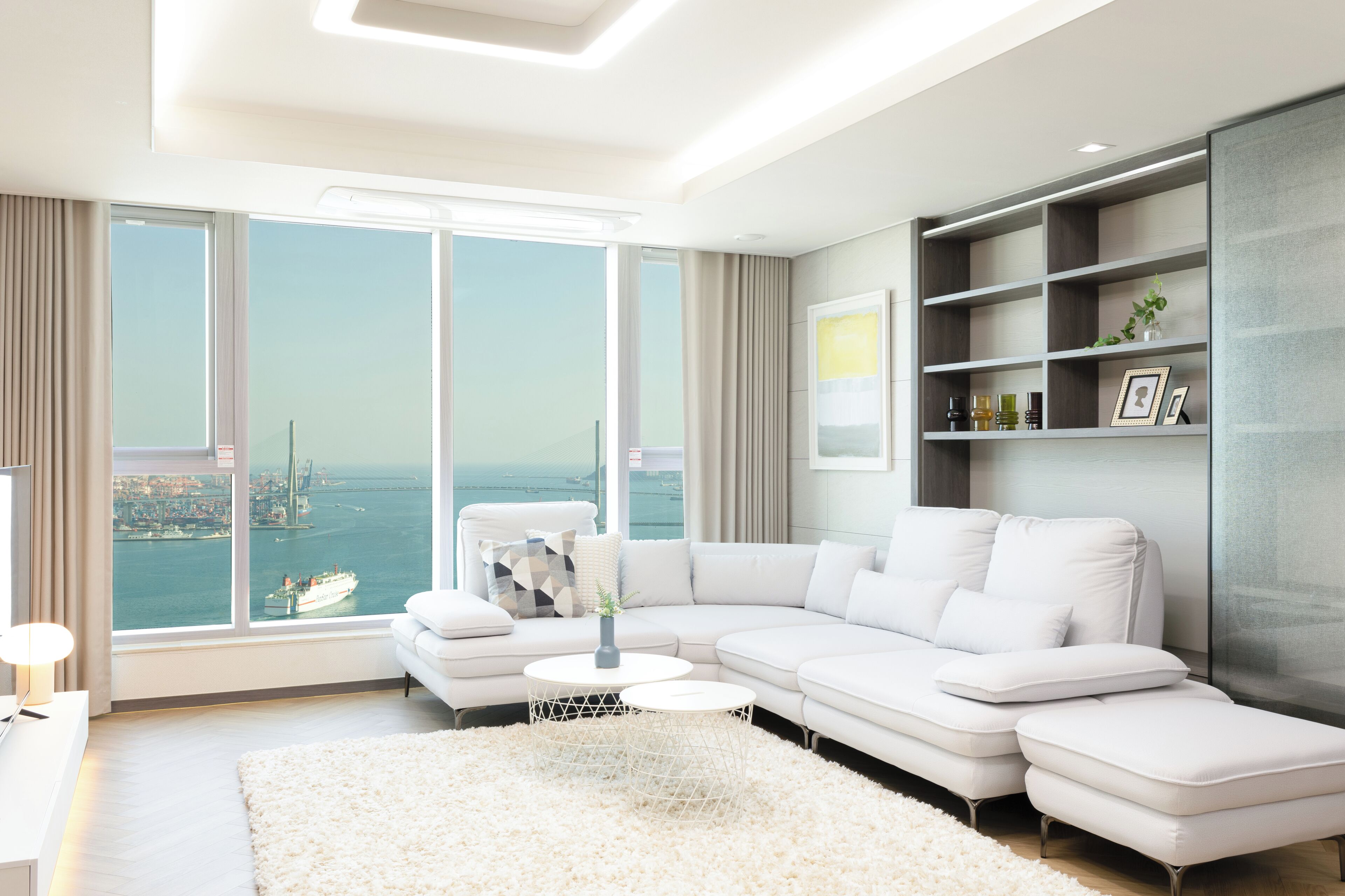 A modern living room with a white sectional sofa, a round coffee table, and a large window offering a city view.