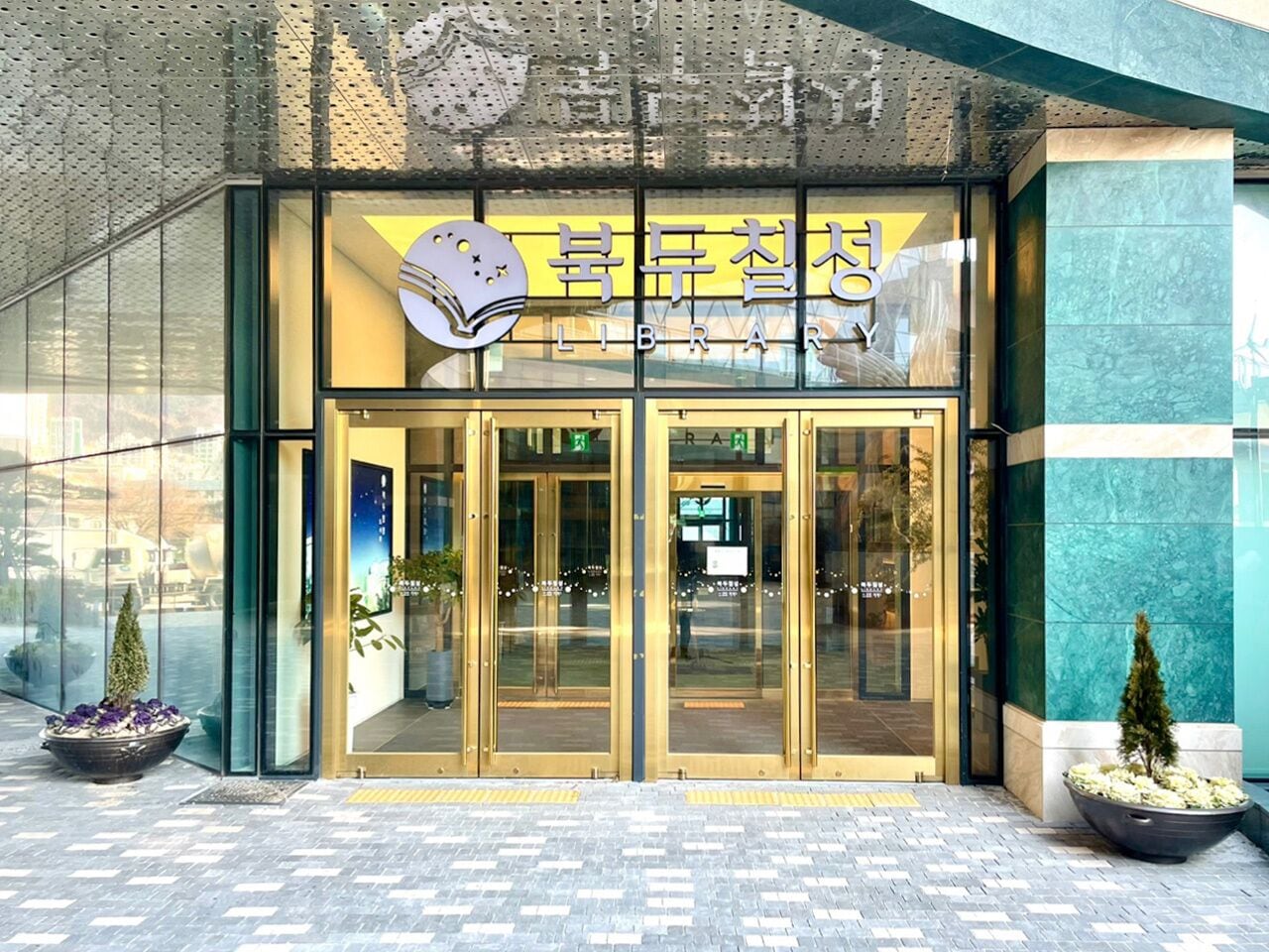 A modern building entrance with glass doors, a sign above reading "LIBRARY," and potted plants on either side.