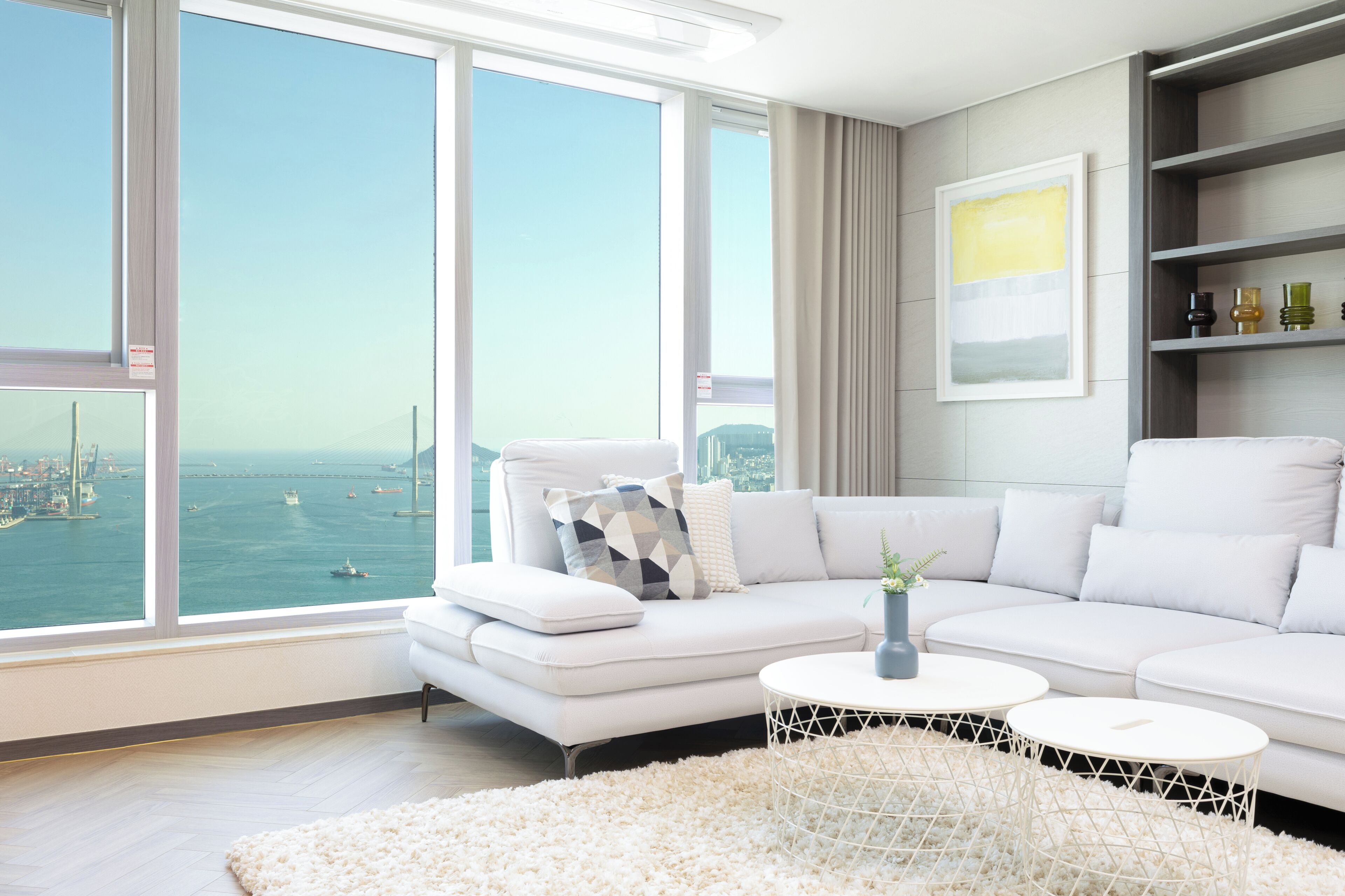 Ein modernes Wohnzimmer mit einem weißen Sofa, zwei runden Couchtischen und Blick auf das Meer sowie die Stadtlandschaft durch große Fenster.