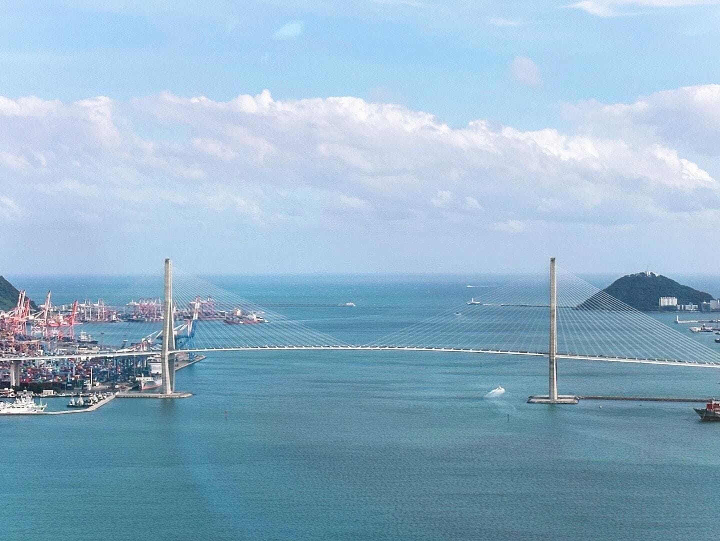 Eine lange Hängebrücke überspannt ein Gewässer, im Hintergrund sind Industriegebiete und eine vorgelagerte Insel zu sehen.