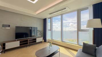 A modern living room with a flat-screen TV, a sofa, a coffee table, and a large window offering a view of a bridge and water.