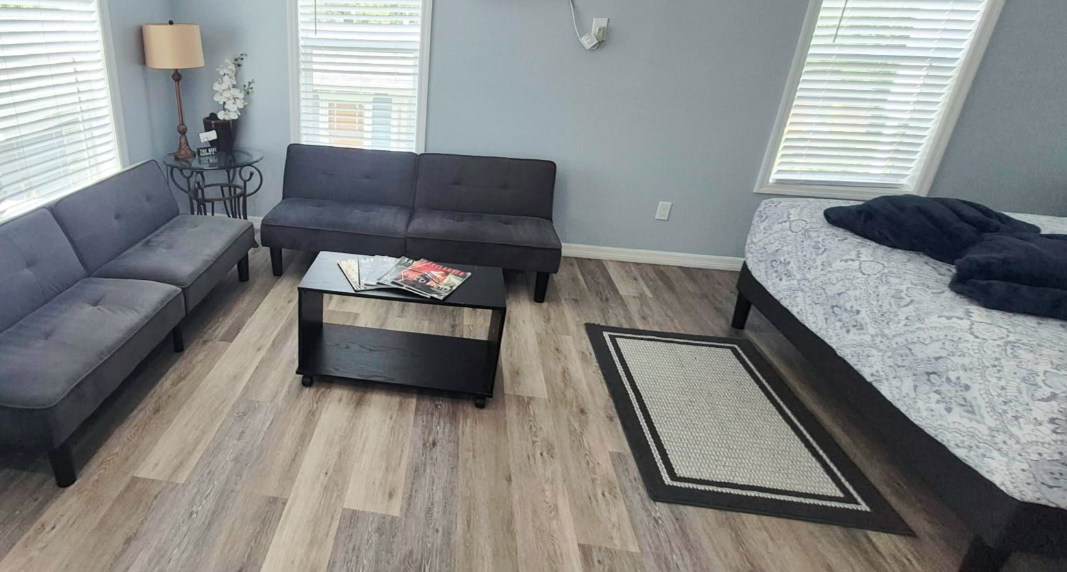 Une pièce avec un canapé gris, une table basse noire sur laquelle sont posés des magazines, une couvre-lit à motifs et une petite table avec une lampe et des fleurs.