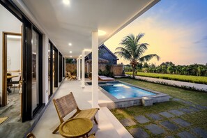 A modern outdoor area with a swimming pool, lounge chairs, and a view of a wooden house and palm trees. - Villa Grateful by Alfred in Bali (Sukawati)