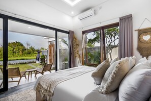 A modern bedroom with a large bed, a balcony with chairs, and a view of greenery. - Villa Grateful by Alfred in Bali (Sukawati)