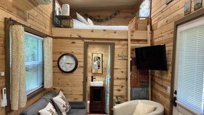 A cozy cabin interior with a sofa, armchair, wooden walls, a clock, a television, and a small kitchen area. - Charming Tiny House on 10 Wooded Acres (Richmond)