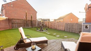 A backyard with artificial grass, a wooden deck, a lounge chair, a small table with a lantern, and a dog in the distance. - Manchester - House - (Manchester)