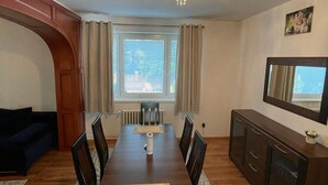 A dining area with a long table, chairs, a radiator, a window with curtains, a dark-colored sofa, and a cabinet with drawers. - 4-Bedroom House Sleeps 7 with Garden (Senec)