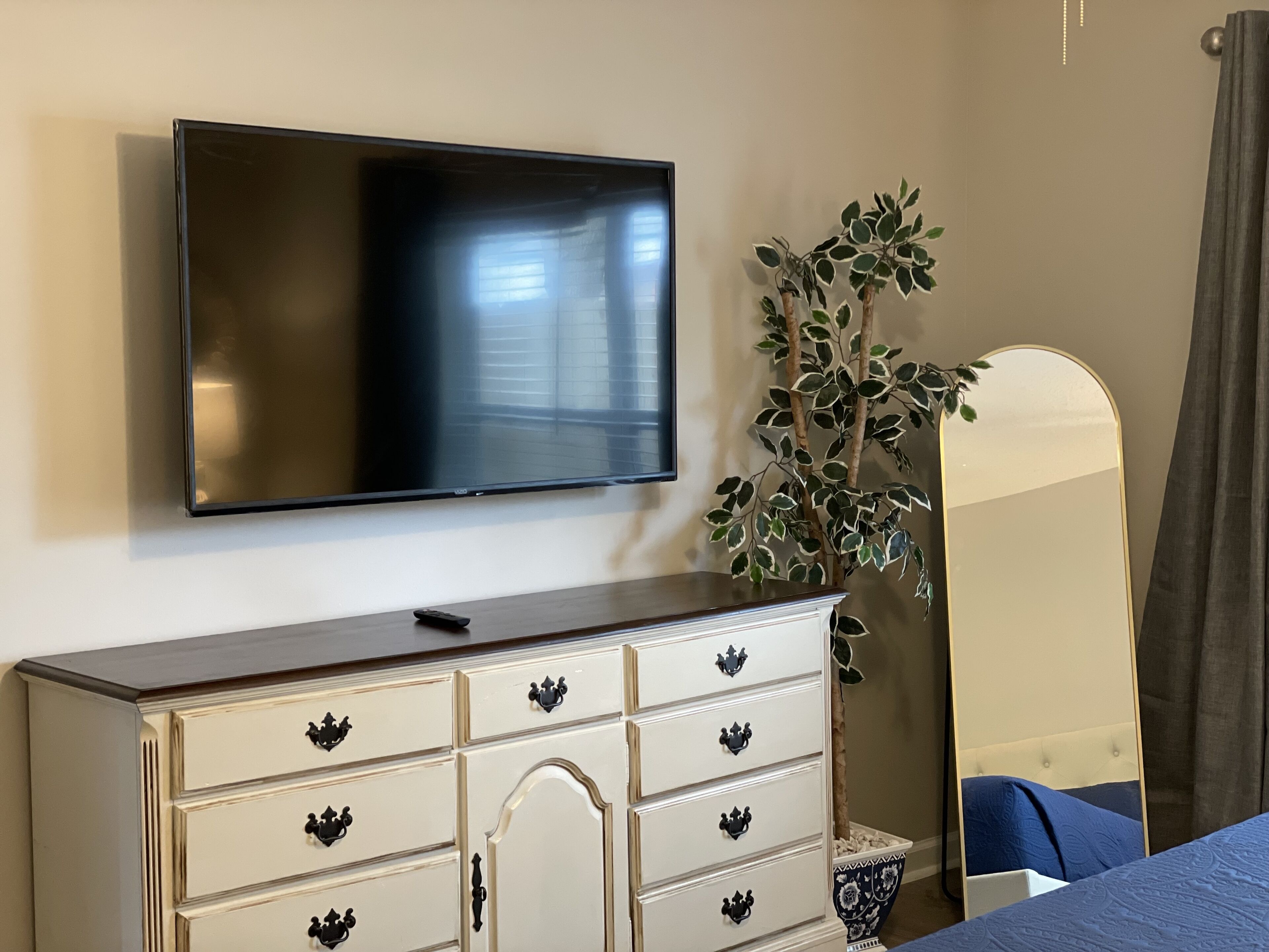 Un dormitorio con una televisión de pantalla plana montada en la pared, un aparador con varios cajones, un espejo y una planta en maceta.