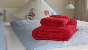 Ein ordentlich bezogenes Bett mit einem blau-weißen Kopfkissen, einem roten Handtuch und einer weißen Lampe im Hintergrund.