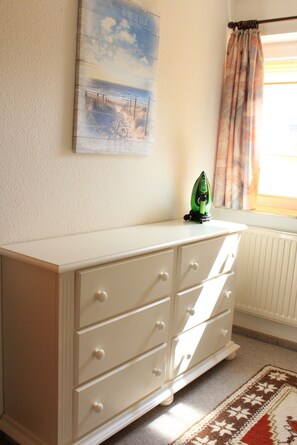 A white dresser with drawers, a green iron, a painting of a beach scene, and a window with curtains.