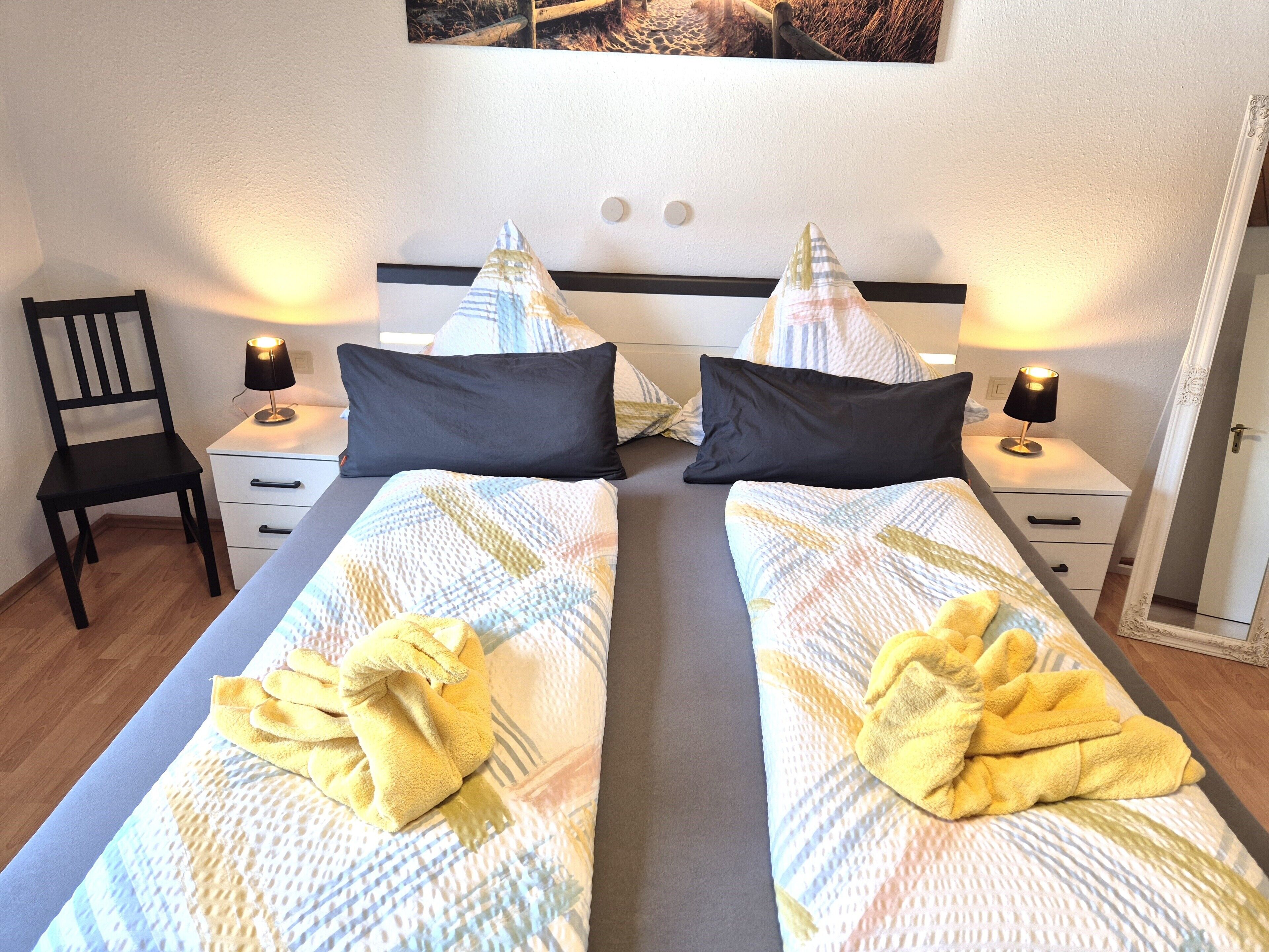 A double bed with a patterned duvet, two black pillows, and a yellow towel on each bed. There are two bedside tables with lamps, a chair, and a mirror on the wall.