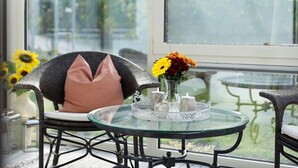Una mesa de cristal con un florero, una silla de mimbre con un cojín y vistas a través de una ventana.