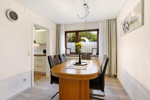 A dining area with a round table, chairs, a vase with flowers, a clock, and a window with curtains.