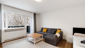 Een moderne woonkamer met een bank, een salontafel, een bed en een raam met uitzicht op kale bomen.