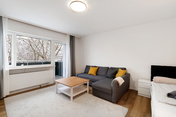 A modern living room with a sofa, a coffee table, a bed, and a window with a view of bare trees.