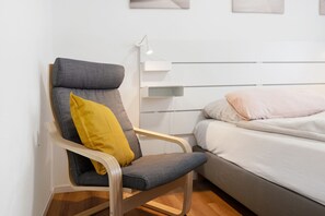 A modern room with a grey upholstered chair, a yellow pillow, a white bedside table, and a bed with white bedding.