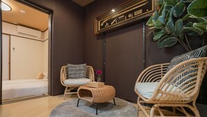 Une pièce confortable avec un lit, deux fauteuils en osier, une petite table sur laquelle est posé un vase, et une grande plante.